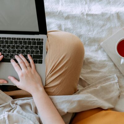 Top view anonymous remote worker typing on keyboard of laptop with blank screen while sitting with crossed legs on bed with tea aside at home